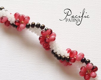 May include: A close-up of a beaded necklace with alternating white and pink beads, forming a flower pattern. The beads are strung on a brown thread. The necklace is on a white surface. The text "Pacific Patina" is visible in the background.
