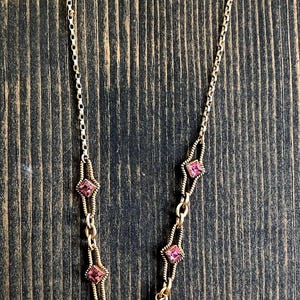 May include: Gold-toned necklace featuring pink gemstones. The necklace has a delicate chain with diamond-shaped and oval-shaped pink stones. The gemstones are set in gold-colored metal. The necklace is displayed on a dark wood surface.