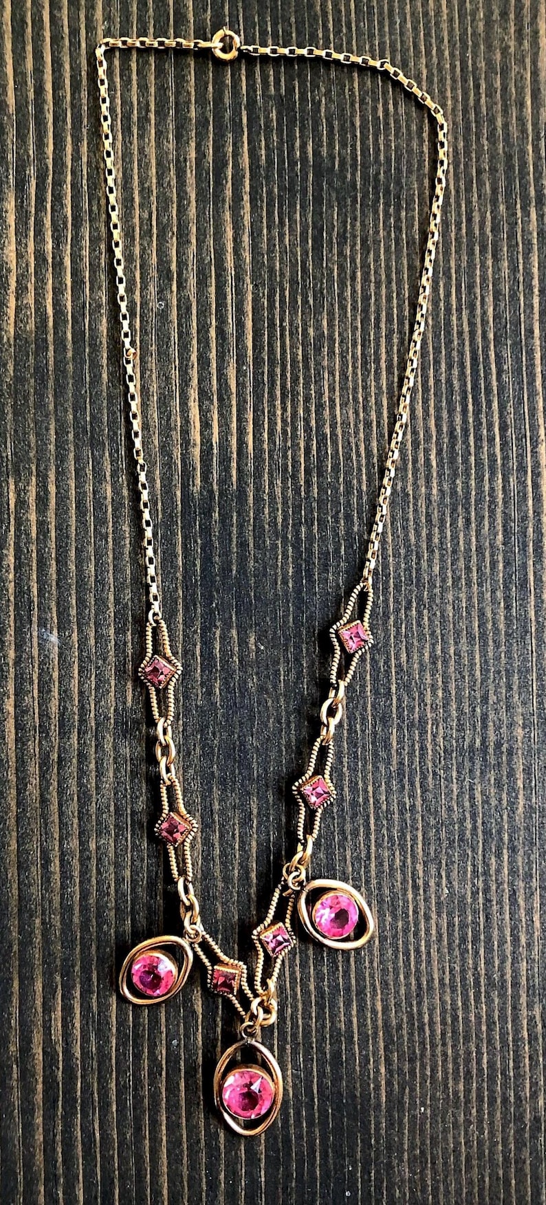 May include: Gold-toned necklace featuring pink gemstones. The necklace has a delicate chain with diamond-shaped and oval-shaped pink stones. The gemstones are set in gold-colored metal. The necklace is displayed on a dark wood surface.