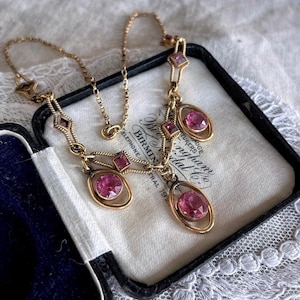 May include: A vintage gold-tone necklace with pink gemstones. The necklace features a delicate chain and is adorned with oval and square-shaped pink stones. The necklace is displayed in a black box with a white interior, showcasing the jewelry's elegance.