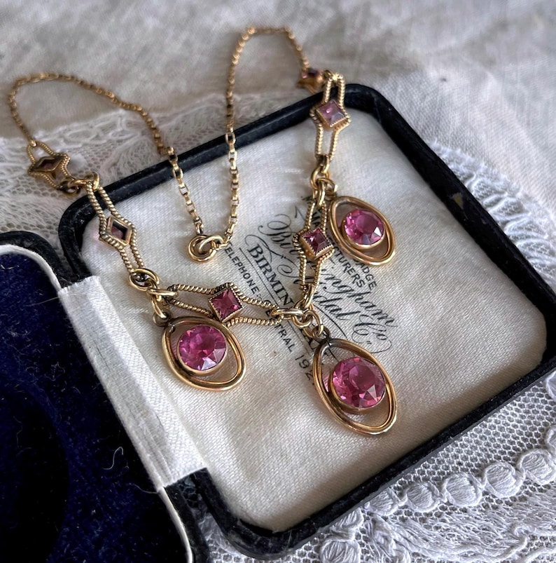 May include: A vintage gold-tone necklace with pink gemstones. The necklace features a delicate chain and is adorned with oval and square-shaped pink stones. The necklace is displayed in a black box with a white interior, showcasing the jewelry's elegance.