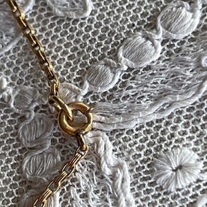 May include: Close-up of a gold-colored necklace chain with a circular clasp. The chain is made of small, rectangular links. The background is a white, textured fabric with a floral pattern.