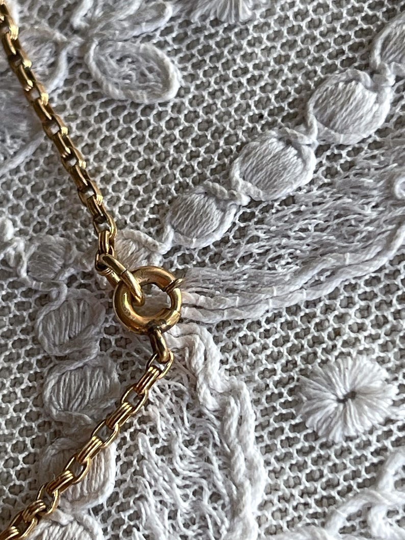 May include: Close-up of a gold-colored necklace chain with a circular clasp. The chain is made of small, rectangular links. The background is a white, textured fabric with a floral pattern.