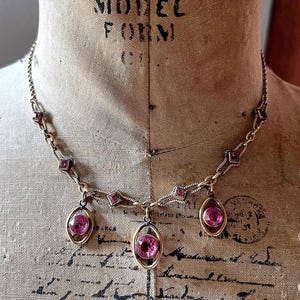 May include: A vintage necklace with a gold-toned chain and setting. The necklace features three oval-shaped pink gemstones suspended from the chain. Additional pink square-shaped gemstones are set along the chain. The necklace is displayed on a vintage mannequin.