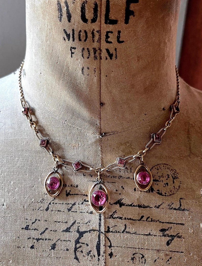May include: A vintage necklace with a gold-toned chain and setting. The necklace features three oval-shaped pink gemstones suspended from the chain. Additional pink square-shaped gemstones are set along the chain. The necklace is displayed on a vintage mannequin.