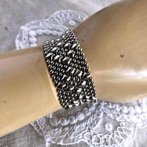 May include: A wide silver-toned bracelet featuring a dense, multi-row mesh design composed of small, reflective metallic beads. The bracelet has a uniform, textured surface with a high-shine finish that catches the light across its entire width.
