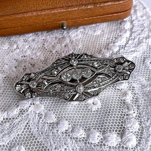 Op de afbeelding: Een antieke zilveren broche met ingewikkelde filigraan en sprankelende, heldere edelstenen. De broche heeft een langwerpig, symmetrisch ontwerp met een centrale cluster van grotere stenen. Het ligt op een delicaat wit kanten oppervlak.