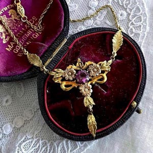 May include: A gold-toned necklace featuring a central purple gemstone surrounded by metal floral and leaf motifs. The pendant is suspended from a delicate chain and rests inside a dark oval jewelry box with a deep red velvet interior lining.