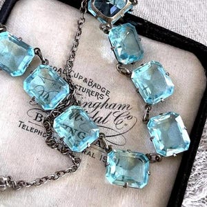 May include: A vintage necklace featuring rectangular, light blue gemstones set in a silver-toned metal. The necklace is displayed on a cream-colored surface with text from "Dal Cup & Badge Manufacturers" and "Bingham Lab Co."