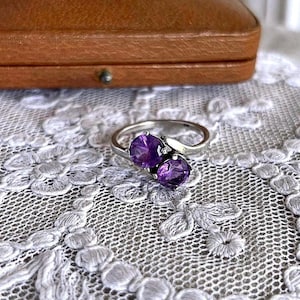 Op de afbeelding: Een zilveren ring met twee ovale, dieppaarse edelstenen. De ring is geplaatst tegen een witte kanten achtergrond, met een bruine leren sieradendoos op de achtergrond.