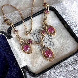 May include: A vintage gold-toned necklace with pink gemstones. The necklace features a delicate chain and pendant design with oval and square-shaped pink stones. The necklace is displayed in a black jewelry box with a white interior.