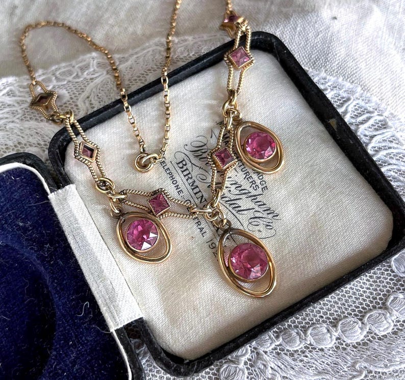 May include: A vintage gold-toned necklace with pink gemstones. The necklace features a delicate chain and pendant design with oval and square-shaped pink stones. The necklace is displayed in a black jewelry box with a white interior.