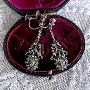 Può includere: Un paio di orecchini pendenti vintage color argento con pietre chiare e scintillanti. Gli orecchini presentano un gruppo di pietre di forma ovale nella parte inferiore, con un motivo floreale sopra. Sono esposti in un portagioie nero con interno in velluto rosso scuro.