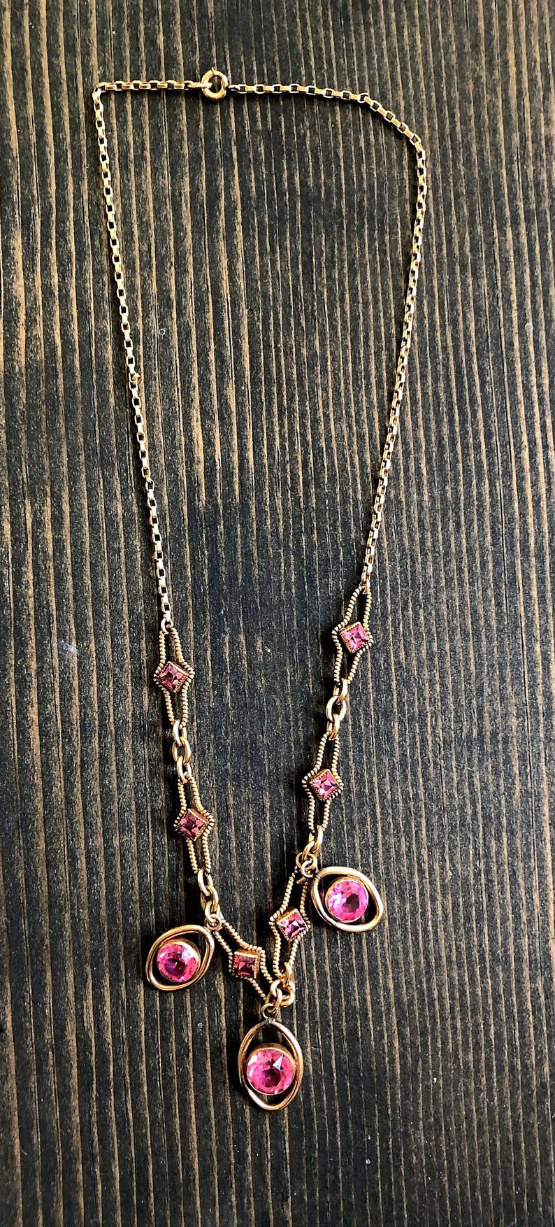 May include: A gold-toned necklace with pink gemstones. The necklace features a delicate chain with alternating pink square and oval-shaped stones. The gemstones are set in gold-colored metal, creating a vintage aesthetic.
