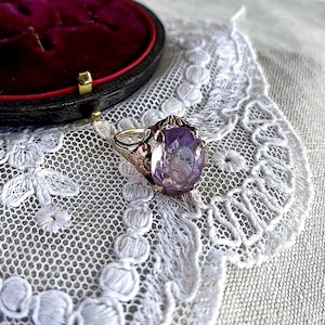 May include: A vintage gold ring with a large, oval-cut amethyst gemstone. The ring is set on a delicate, ornate band. The ring is displayed on a white lace doily, with a burgundy jewelry box in the background.