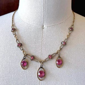 May include: A vintage gold-toned necklace featuring pink gemstones. The necklace has a delicate chain with square and oval-shaped settings. Three oval pendants with large pink stones hang from the center.