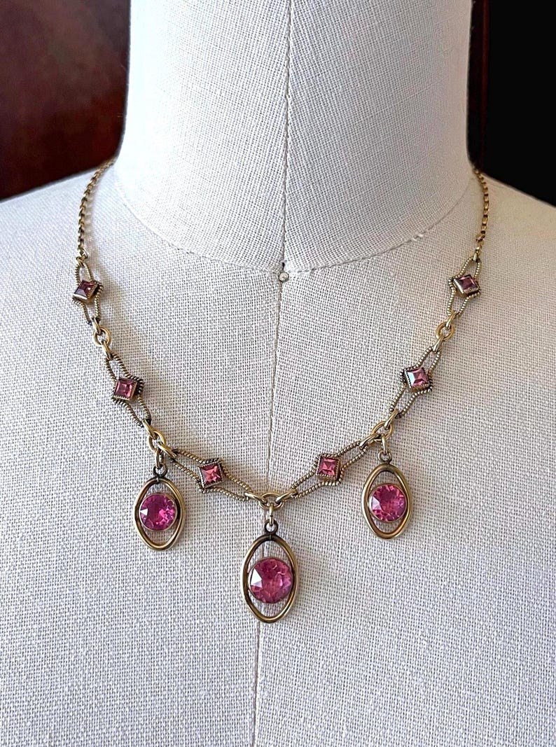 May include: A vintage gold-toned necklace featuring pink gemstones. The necklace has a delicate chain with square and oval-shaped settings. Three oval pendants with large pink stones hang from the center.