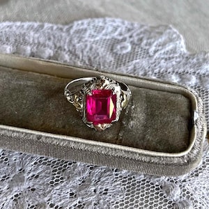 May include: A vintage ring with a large, rectangular, bright pink gemstone set in a silver and gold filigree setting. The ring is displayed in a gray velvet ring box, set against a backdrop of white lace.