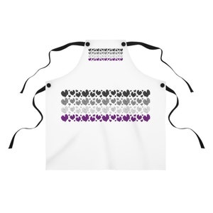 Puede incluir: Un delantal blanco con correas ajustables negras y un diseño de bandera del orgullo asexual hecha de corazones en tonos de gris, negro y morado.