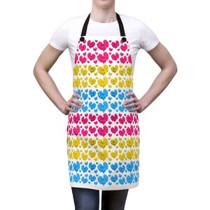 Puede incluir: Un delantal blanco con un patrón de corazones rosas, amarillos y azules. El delantal tiene tirantes ajustables y un bolsillo.