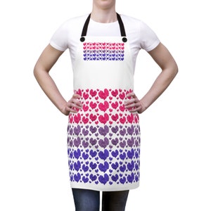 Puede incluir: Un delantal blanco con un patrón de corazones rosa, morado y azul. Los corazones están dispuestos en un patrón de degradado, con rosa en la parte superior y azul en la parte inferior. El delantal tiene correas ajustables y un bolsillo.