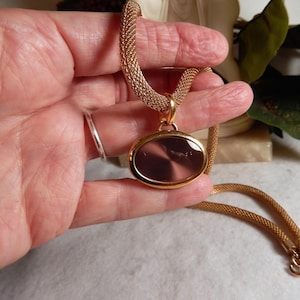 Peut inclure: Collier doré avec un pendentif ovale au centre translucide foncé. Le pendentif est encadré d'or et suspendu à une chaîne en maille. Le collier est présenté, mettant en valeur son design et sa couleur.