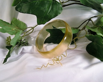 Könnte beinhalten: Ein hellbraunes Armreif-Armband mit glatter, glänzender Oberfläche. Das Armband liegt auf einer weißen Oberfläche mit grünen Blättern.