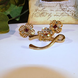 May include: A gold-toned brooch with two flower-shaped charms. Each flower has a red rhinestone center and clear rhinestones around the center. The brooch is in the shape of a curved line.