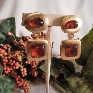 May include: Gold-toned clip-on earrings with amber-colored glass inserts. Each earring features a round top and a square bottom, connected by a small hinge. The earrings are displayed on a white stand, with greenery and berries in the background.