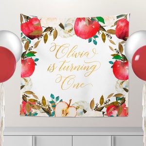 May include: A white backdrop with a gold inscription that reads "Olivia is turning One". The backdrop is decorated with a watercolor border of red apples, green leaves, and white flowers.