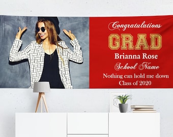 May include: Red and gold graduation banner with the text "Congratulations Grad" and the name "Brianna Rose". The banner also includes the text "School Name", "Nothing can hold me down", and "Class of 2020". A photo of a woman in a black and white patterned jacket is on the left side of the banner.