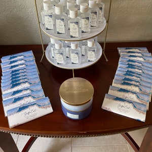 May include: A gold-coloured, two-tiered serving stand displays numerous small, clear bottles of hand sanitiser with white tops. The bottles have labels with a cross and text. Several stacks of blue and white cards and a blue and gold candle are also present.