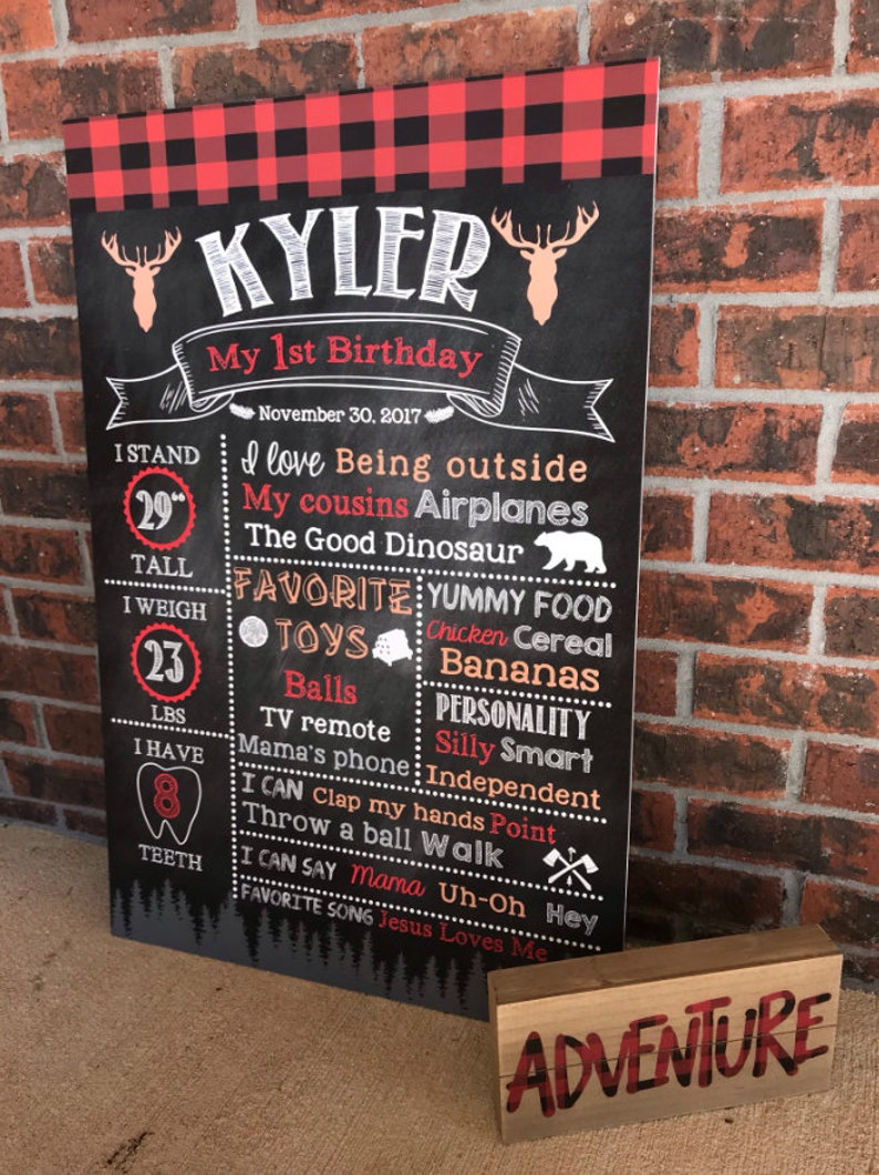 May include: A chalkboard-style sign for a first birthday, featuring the name "Kyler" and details like height (29 inches), weight (23 lbs), and favorite things. A wooden block with "ADVENTURE" is in front.