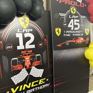 May include: Two black and red cardboard signs with a Formula 1 racing theme. The signs feature a red race car, checkered flags, and the text "LAP 12" and "LAP 45" with the words "VINCE HAPPY BIRTHDAY" and "PAOLO * AROUND * THE TRACK" on the signs.
