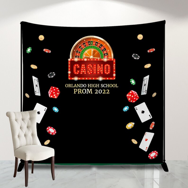 May include: A black backdrop with casino-themed graphics, including a roulette wheel, playing cards, dice, and poker chips. The word "CASINO" is displayed in red neon lights, with "ORLANDO HIGH SCHOOL PROM 2022" below. A white upholstered chair is in the foreground.
