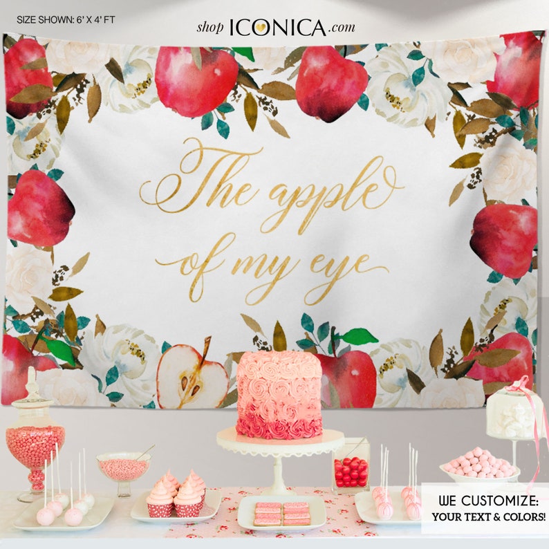 May include: A white backdrop with a floral border featuring red apples and white flowers. The text "The apple of my eye" is written in gold script. The backdrop is 6 feet by 4 feet.