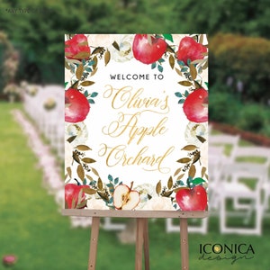 May include: Welcome to Olivia's Apple Orchard sign with a white background and a wreath of red apples, white flowers, and green leaves. The text is in gold script.