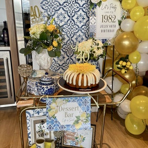 Op de afbeelding: Een gouden barwagen versierd met een blauw-witte tegelpatroon. De wagen is gestyled voor een 40e verjaardagsfeest met een taart, ballonnen en borden met de tekst "Let's Party Like It's 1982" en "Dessert Bar".