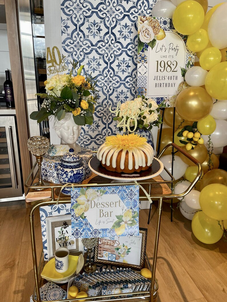 Op de afbeelding: Een gouden barwagen versierd met een blauw-witte tegelpatroon. De wagen is gestyled voor een 40e verjaardagsfeest met een taart, ballonnen en borden met de tekst "Let's Party Like It's 1982" en "Dessert Bar".