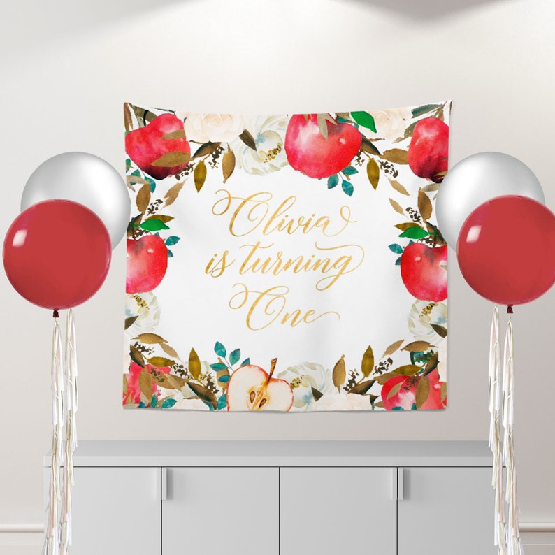May include: A white backdrop with a gold inscription that reads "Olivia is turning One". The backdrop is decorated with a watercolor border of red apples, green leaves, and white flowers.
