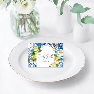Op de afbeelding: Een wit plaatskaartje met een blauw en geel betegeld patroon en een citroentak ontwerp. De tekst op het kaartje luidt "Emily Smith, Tafel 1."