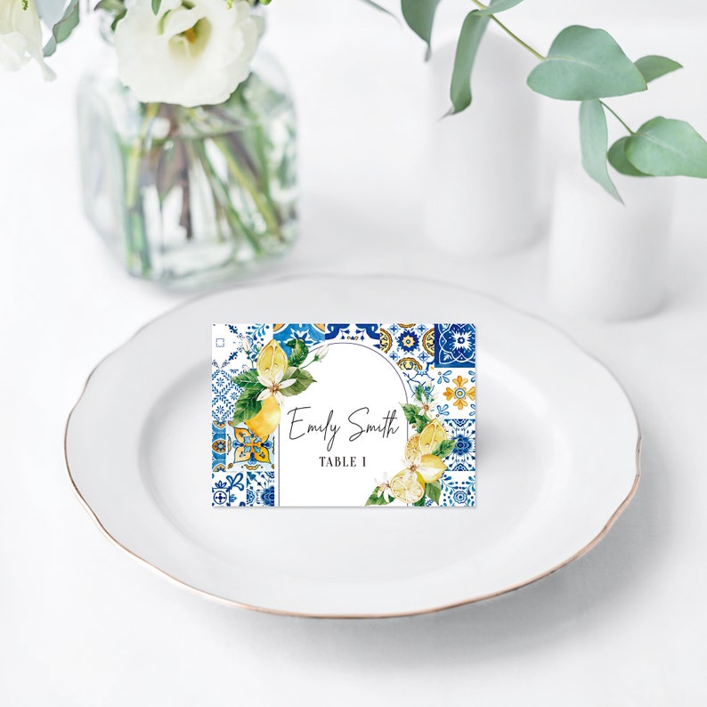 Op de afbeelding: Een wit plaatskaartje met een blauw en geel betegeld patroon en een citroentak ontwerp. De tekst op het kaartje luidt "Emily Smith, Tafel 1."