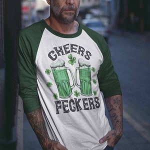 May include: A white and green baseball style t-shirt with a green and white graphic of two beer mugs with shamrocks and the text "Cheers Feckers".
