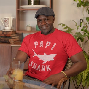 May include: A man wearing a red t-shirt with a white shark graphic and the text "Papi Shark". He is wearing a grey newsboy cap and has a glass of orange juice in his hand.