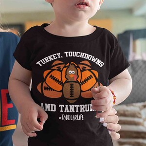 May include: A black toddler t-shirt with a graphic of a turkey made of footballs. The text on the shirt reads "Turkey, Touchdowns and Tantrums #ToddlerLife".