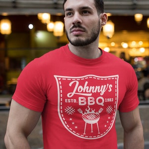 May include: A red t-shirt with a white graphic that reads "Johnny's BBQ Estd. 1983". The graphic features a shield design with a grill, stars, and crossed lines. The shirt is short-sleeved.