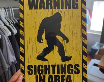 May include: Yellow warning sign with black stripes and text. The sign reads "WARNING SIGHTINGS IN AREA". A black silhouette of Bigfoot is walking across the sign.