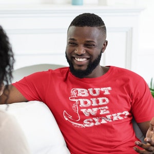 May include: A man and a woman are sitting on a white couch. The man is wearing a red t-shirt with the text "But Did We Sink?" and a white anchor graphic.
