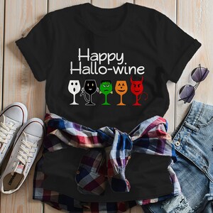 K&ouml;nnte beinhalten: Schwarzes T-Shirt mit dem Text "Happy Hallo-wine" und einer Reihe von f&uuml;nf Weingl&auml;sern mit verschiedenen Halloween-Charaktergesichtern.