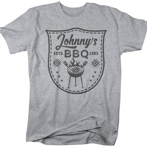 May include: A gray t-shirt featuring a shield-shaped graphic with the text "Johnny's BBQ ESTD. 1983." The graphic includes a grill, flames, and stars. The shirt is a casual wear item.